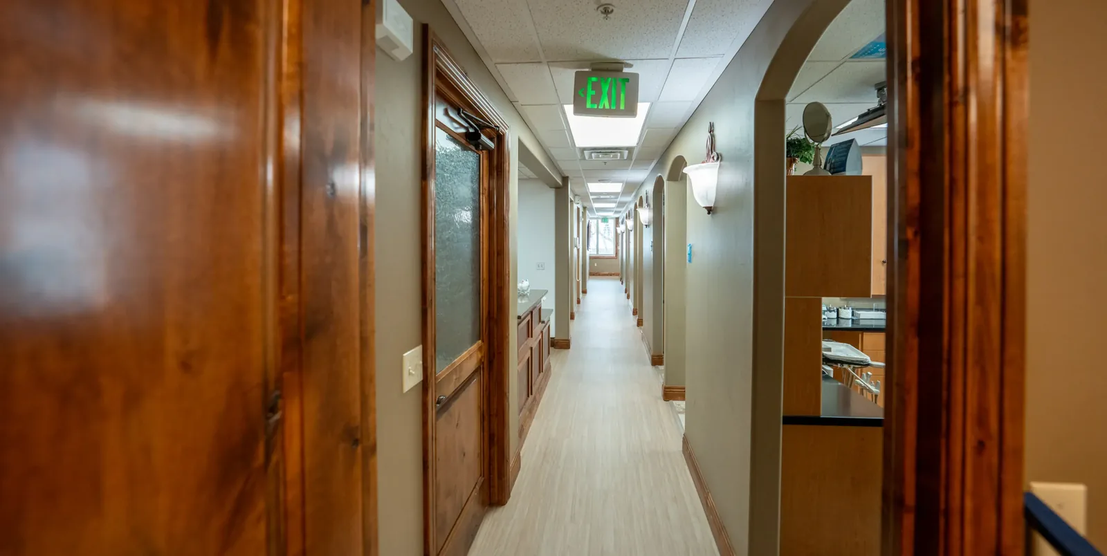Hallway in a dental clinic with wooden doors, arched treatment room entrances, light flooring, and an EXIT sign.