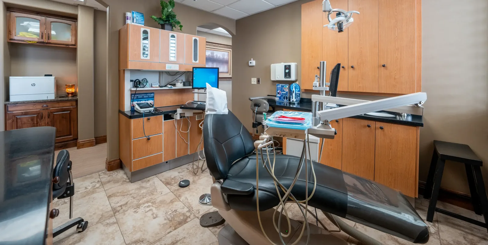 Dental examination room featuring a black patient chair, overhead light, instruments, and wooden cabinets.